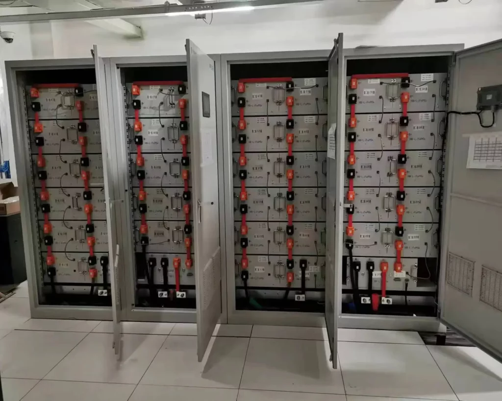 A row of industrial and commercial energy storage cabinets have their doors open, with multiple battery modules connected in series and parallel to form a battery cluster, as well as some electronic control devices such as PCS and EMS. These devices are labeled with text and small display screens. The cabinets are located in a clean industrial environment.