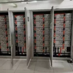 A row of industrial and commercial energy storage cabinets have their doors open, with multiple battery modules connected in series and parallel to form a battery cluster, as well as some electronic control devices such as PCS and EMS. These devices are labeled with text and small display screens. The cabinets are located in a clean industrial environment.