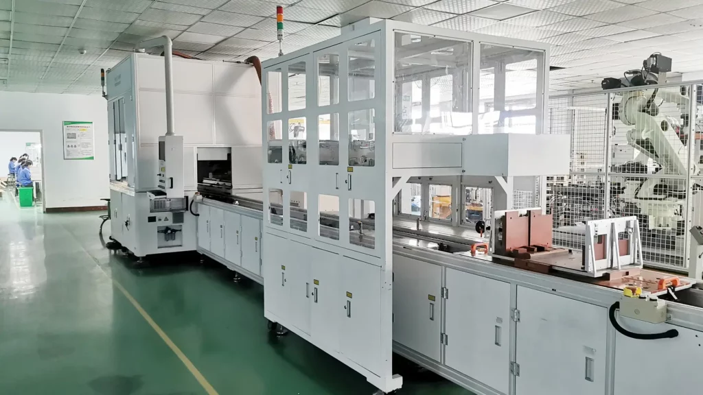 A battery pack production line with conveyor belts in a modern manufacturing plant with automated machinery. In the background are other machines and several workers in blue uniforms.